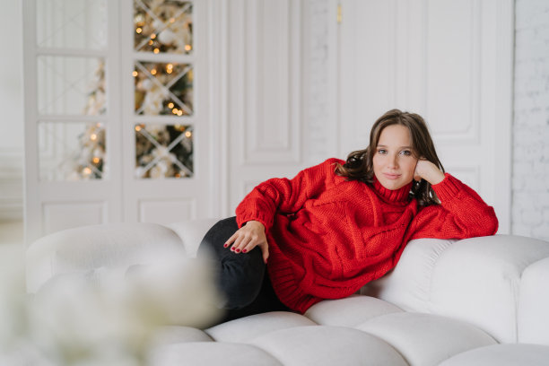 照片上的年轻女子穿着温暖的针织衣服，躺在舒适的沙发上，享受着家庭气氛，背景是装饰好的新年树。人，休闲和业余时间的概念图片下载