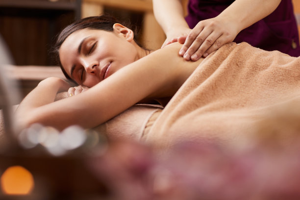 满意的年轻女子棕色头发躺在按摩台上闭着眼睛，而专业按摩师揉她的上背部图片下载