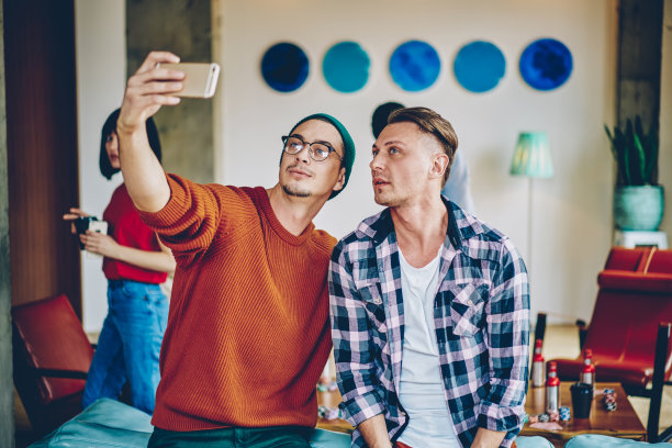 两个时尚的穿着休闲的潮男看着智能手机的前置摄像头，在社交网络上发布自拍照片。年轻人在友好的会议期间为博客拍摄视频图片下载
