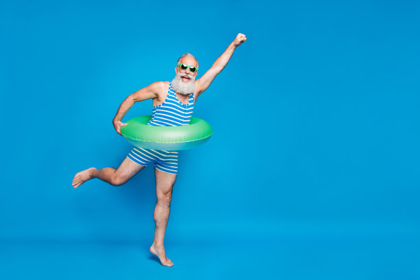 完整的照片兴奋的退休老人与灰色的头发举起他的拳头举着玩具圈，穿着条纹泳衣眼镜孤立在蓝色的背景图片下载