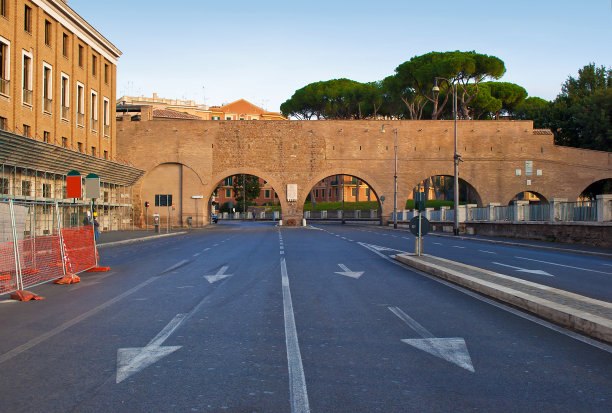一个宽阔空旷的道路与白色的线和箭头和路灯到一个拱形砖墙的形象。温暖的早晨，秋高气爽，万里无云图片下载