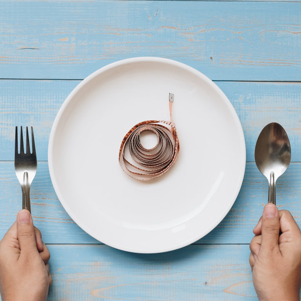 俯视图女人手里拿着勺子和叉子，上面是白色的盘子和粉红色的卷尺，放在蓝色的木桌上。节食、减肥和清洁食品的概念图片下载