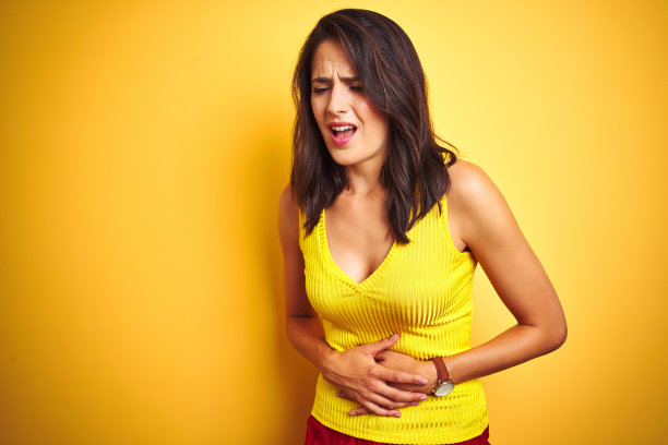 年轻美丽的女子穿着t恤站在黄色孤立的背景与手胃，因为消化不良，痛苦的疾病感觉不舒服。疼痛的概念。图片下载
