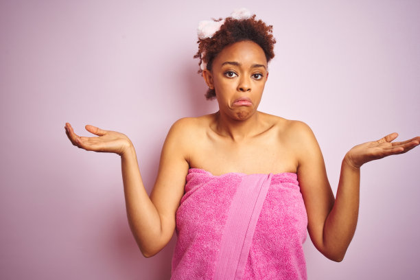 非洲裔美国妇女穿着淋浴毛巾后，在粉红色孤立的背景愚蠢和困惑的表情与胳膊和手举起。怀疑的概念。图片下载
