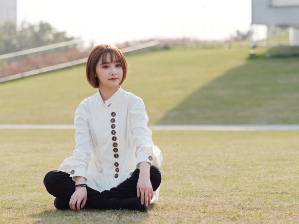 美丽的年轻女子在白色衬衫与腿交叉坐在草地和微笑的相机在阳光灿烂的日子。户外时尚写真魅力中国开朗时尚女孩。图片下载