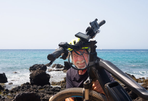 一位白发苍苍、身后是蓝色海洋的老人的照片很不寻常。享受自行车运动的健康生活方式。与波的海岸线。戴着黄色头盔的快乐男子在阳光下享受电动自行车图片下载