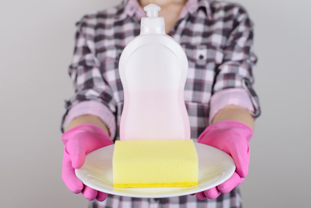 瓶相机清布推广女佣脏盘子人家庭晚餐人的概念。近距离的照片女士的手拿着瓶子黄色海绵白色盘子在手上孤立的灰色背景图片下载