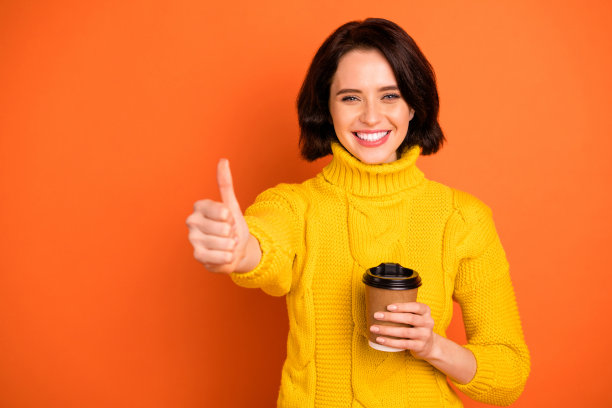 照片的愉快的好可爱迷人的漂亮甜美的女孩牙齿微笑显示你的拇指向上标志穿着黄色毛衣，而孤立与橙色的背景图片下载