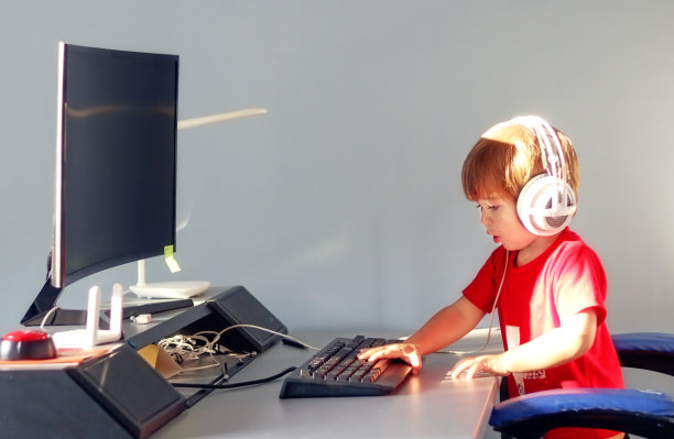 有趣好奇的小男孩戴着耳机在键盘上打字，坐在他哥哥的电脑显示器前。孩子和互联网。现代的童年。图片下载