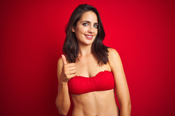 美丽的女人穿着比基尼泳装在红色孤立的背景做快乐的大拇指向上的手势与手。赞许的表情看着镜头，表示成功。图片下载