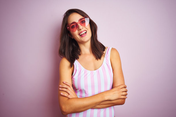 美丽的女人穿着条纹粉色泳衣和心形太阳镜在粉红色的背景快乐的脸微笑着交叉双臂看着镜头。积极的人。图片下载