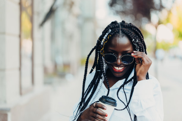 这是一个阳光明媚的户外，一个扎着辫子、皮肤黝黑、戴着眼镜的年轻女孩的肖像，手里拿着咖啡。户外的照片。图片下载