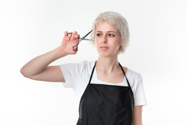 漂亮的年轻发型师在白色t恤和黑色围裙拿着黑色剪刀靠近脸准备工作站在孤立的白色背景。图片下载