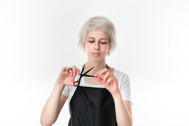 漂亮的年轻发型师在白色t恤和黑色围裙拿着黑色剪刀靠近脸准备工作站在孤立的白色背景。图片下载