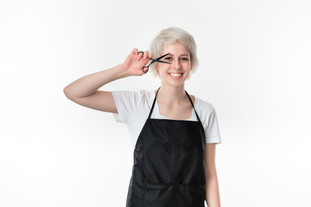 漂亮的年轻发型师在白色t恤和黑色围裙拿着黑色剪刀靠近脸准备工作站在孤立的白色背景。图片下载