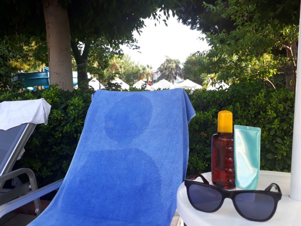 太阳床旁边的桌子上放着防晒油、防晒胶和太阳镜，上面有一条蓝色的毛巾。图片下载