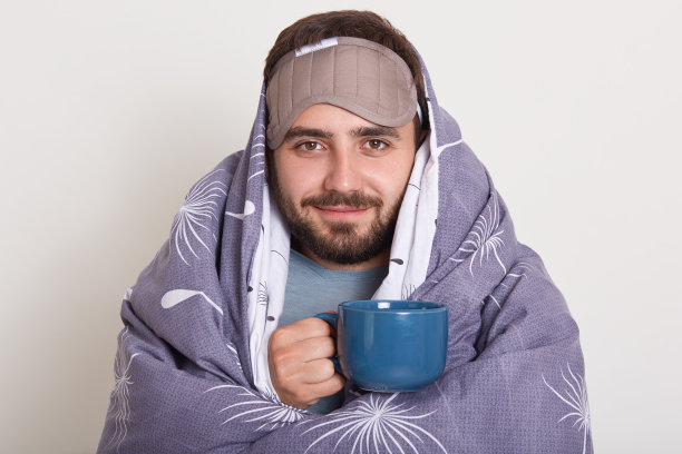 这是一幅前额蒙着眼罩的蓄着胡须的高加索青年男子的照片，他手拿蓝色的咖啡杯或马克杯，裹着灰色的毯子，在白色的画室背景下摆姿势。图片下载