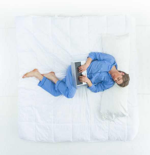 英俊的男人穿着睡衣躺在床上用着他的工作笔记本电脑图片下载