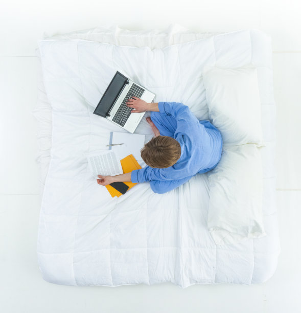 英俊的男人穿着睡衣躺在床上用他的工作笔记本电脑和文件图片下载