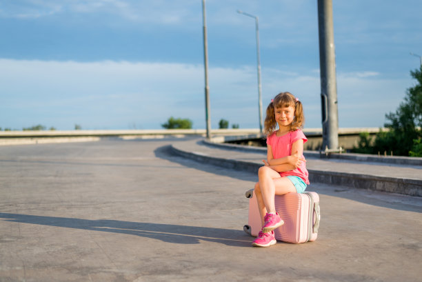 小女孩坐在路边的一个粉色行李箱上，空着。图片下载