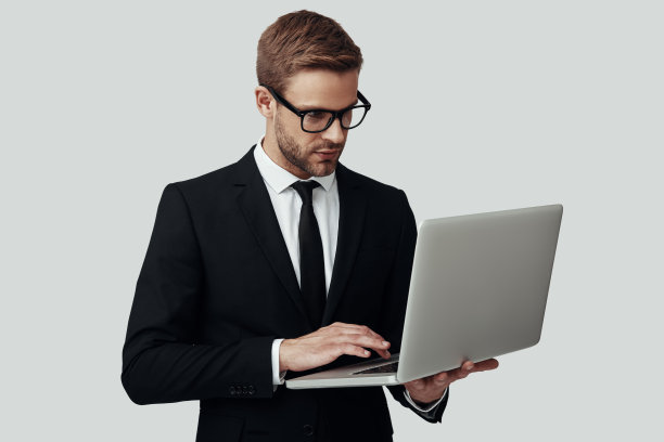 帅气的年轻人穿着正装使用笔记本电脑工作，而站在灰色背景图片下载