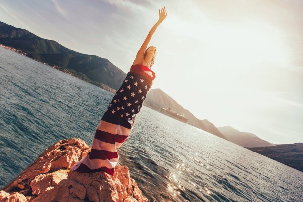 美丽的年轻女子站在礁石上裹着美国国旗。图片下载