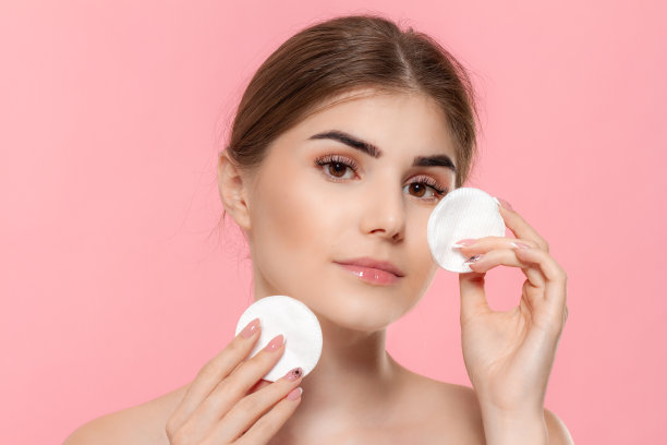 美模用白色的棉海绵把化妆品涂到皮肤上。模型看着孤立的粉红色背景的相机。图片下载