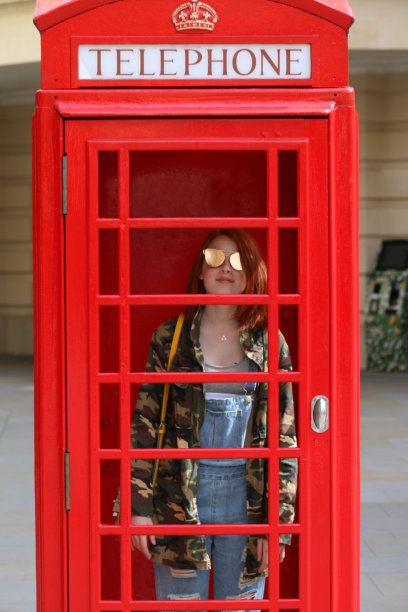 这是一个14 / 15岁的红发少女站在英国街道上一个废弃的红色电话亭里，戴着太阳镜，穿着一件复古的军用迷彩服，外面是蓝色的粗斜纹棉布，肩上挎着一个黄色的手提包图片下载