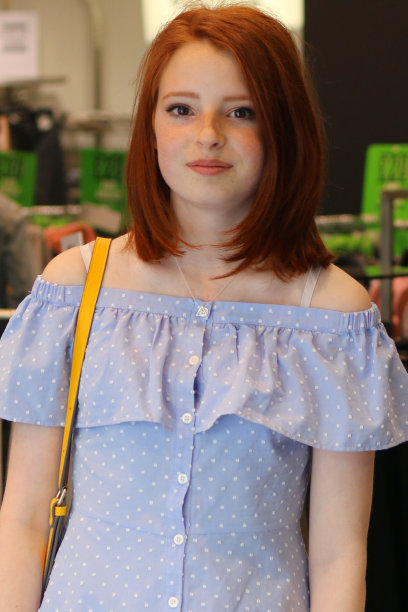 这是一个14 / 15岁的红发少女站在商店里的照片，她穿着一件漂亮的淡蓝色带纽扣的带有白色圆点图案的巴多连衣裙，肩上挎着一个黄色的手提包图片下载