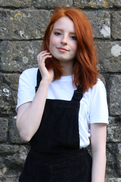 照片中，一个14 / 15岁的红发少女，皮肤苍白，脸上有雀斑，站在一堵长满青苔的旧石墙前，穿着一件时髦休闲的黑色罗纹灯芯绒围裙裙，梳着波浪发型图片下载