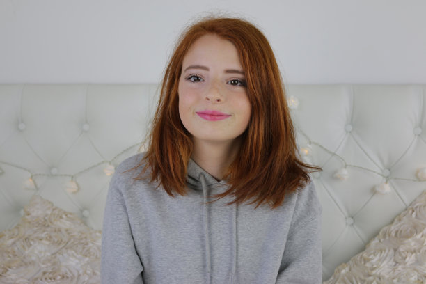 照片中，14 / 15岁的红发少女，皮肤苍白，脸上有雀斑，穿着灰色的兜帽上衣，涂着粉色的口红，坐在卧室里看着镜头，神情轻松，面带微笑图片下载