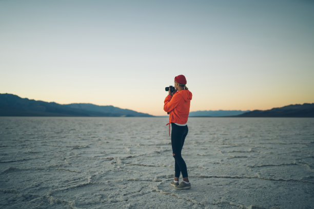 熟练的女摄影师拍摄美丽的黄昏风景在沙漠的风景，年轻的女人在旅行中穿着时髦的橙色夹克拍摄自然照片的背影图片下载