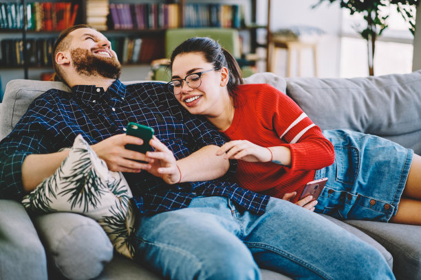 愉快浪漫的情侣在爱笑在有趣的照片上看智能手机一起休息在沙发上，年轻的婚姻有乐趣看视频坐在客厅的沙发上手机图片下载