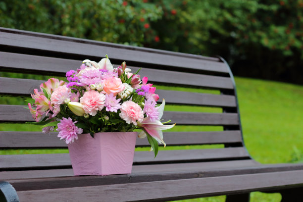 长凳上放着一束鲜花的粉色盒子。粉色，白色和淡紫色，康乃馨，百合，玫瑰和绿叶。被遗忘在公园里的花。室外装饰摄影拍摄，婚礼庆典户外概念图片下载