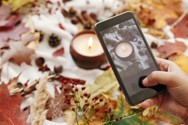 拿着手机，拍着秋平躺着的照片。蜡烛，浆果，落叶，茴香，香草，橡子，坚果，肉桂，白色纺织品上的棉花。舒适的生活方式，惬意的秋天心情图片下载