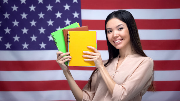亚洲女孩对着美国国旗的背景微笑，学生拿着抄写本图片下载