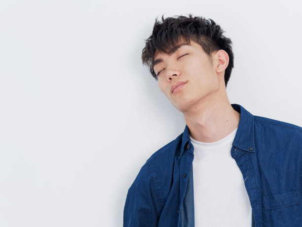 一个穿着蓝色衬衫的英俊的中国年轻男子的肖像靠在墙上，闭着眼睛，疲倦的表情，孤立在白色的背景。图片下载