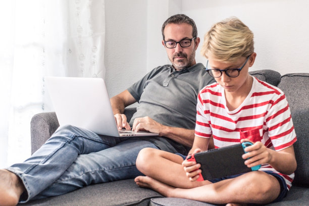 当爸爸坐在沙发上使用笔记本电脑时，小男孩在玩视频游戏机。父亲和儿子，世代相比较。新技术、通讯、生活理念图片下载