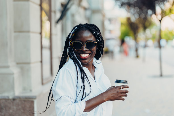 这是一个阳光明媚的户外，一个扎着辫子、皮肤黝黑、戴着眼镜的年轻女孩的肖像，手里拿着咖啡。户外的照片。图片下载