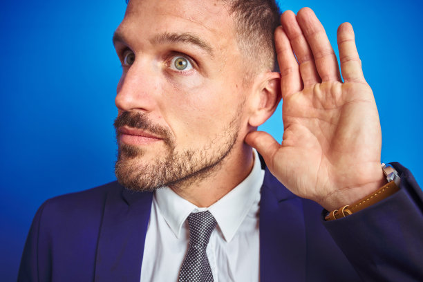近距离图片的年轻英俊的商人在蓝色孤立的背景微笑与手捂耳朵听谣言或八卦。耳聋的概念。图片下载