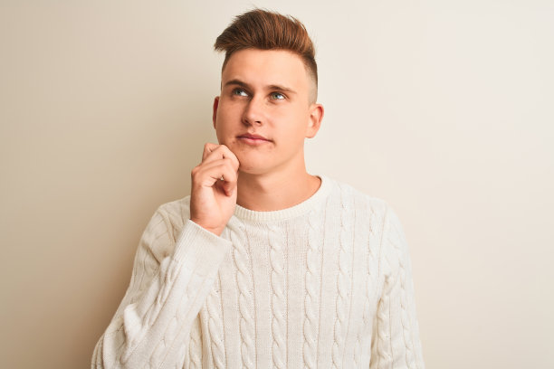 年轻英俊的男子穿着冬季毛衣站在孤立的白色背景与手下巴思考问题，沉思的表情。带着沉思的微笑。怀疑的概念。图片下载
