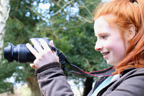 年轻的少女学习摄影用数码单反相机拍照，聚焦镜头在郊外穿着绿色打过蜡的夹克，红色的头发马尾辫/姜黄色的发型，看着相机屏幕的取景器图片下载