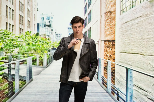腰部以上/一个20-29岁的白人男性/年轻男性谁是站在户外穿着牛仔裤/皮夹克谁是认真的/自信/集中/冷静的态度和拿着咖啡杯/美国图片下载