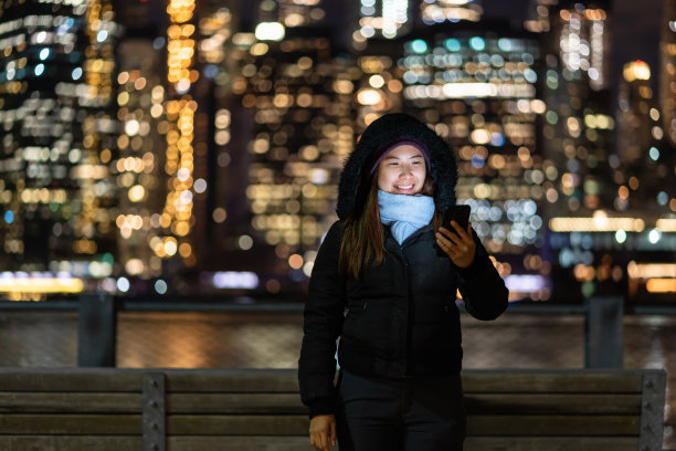 亚洲女人穿着冬季套装用智能手机用微笑动作拍下了模糊的散景照片，旁边是东河的背景，美国市中心，职业理念图片下载