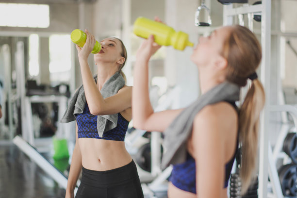 年轻苗条的女人在健身房锻炼后休息，这是喝水。一个女人站在健身房里的大镜子前喝水。从后面看。运动女孩的概念。图片下载