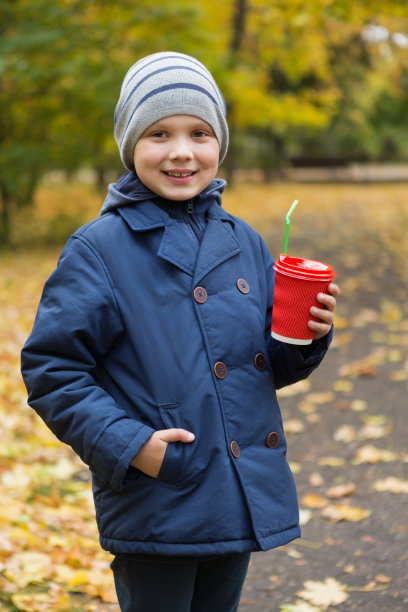 男孩走过秋天的公园，面带微笑，拿着红色的纸板玻璃杯，热饮，秋天的概念图片下载