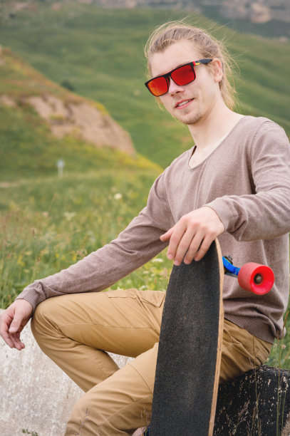 一个戴着墨镜留着长发的年轻时尚男子，手里拿着一块长板坐在乡间柏油路上的削片机上图片下载