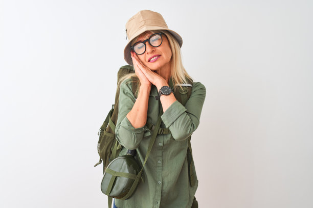 中年徒步妇女戴双肩包帽水壶眼镜在孤立的白色背景睡累了做梦和摆姿势双手同时闭上眼睛微笑。图片下载