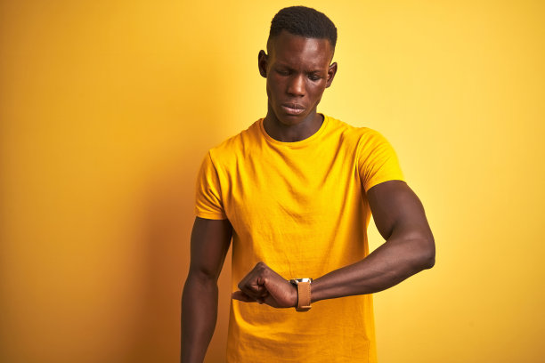 年轻的非洲裔美国人穿着休闲t恤站在孤立的黄色背景上看手表上的时间，放松和自信图片下载