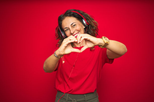中年妇女戴着耳机听音乐在红色孤立的背景微笑在爱显示心形符号和形状的手。浪漫的概念。图片下载